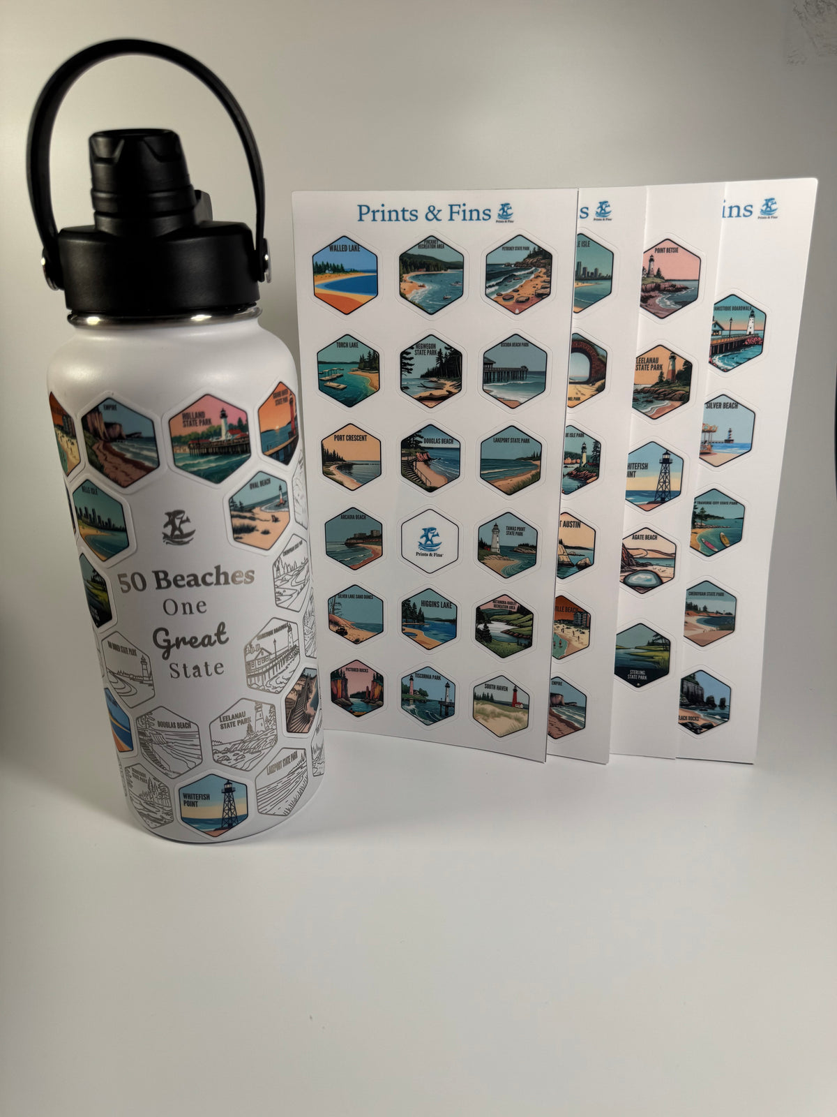 A white insulated water bottle with a black lid and carry loop, printed with beach scene hexagons and the text '50 Beaches One Great State,' shown beside two printed sheets of matching hexagonal beach prints.